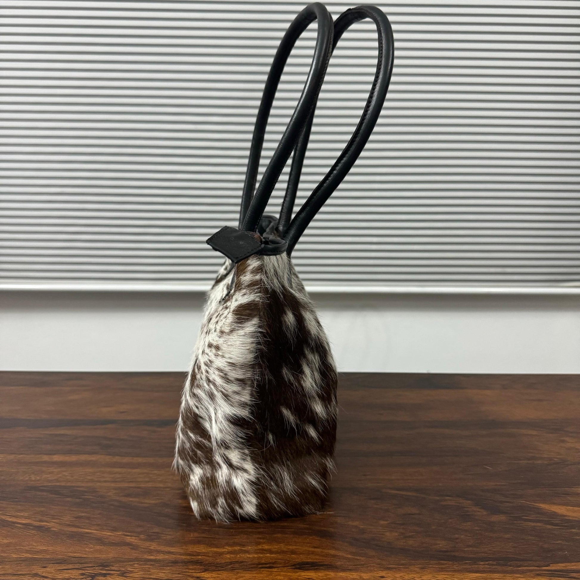 A small handbag with a brown and white cowhide body and black double shoulder straps, standing on a dark wooden surface in front of a gray slatted window.