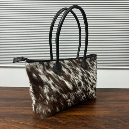 A cowhide handbag with a brown and white spotted pattern and black leather handles, resting on a wooden table against a gray slatted background.