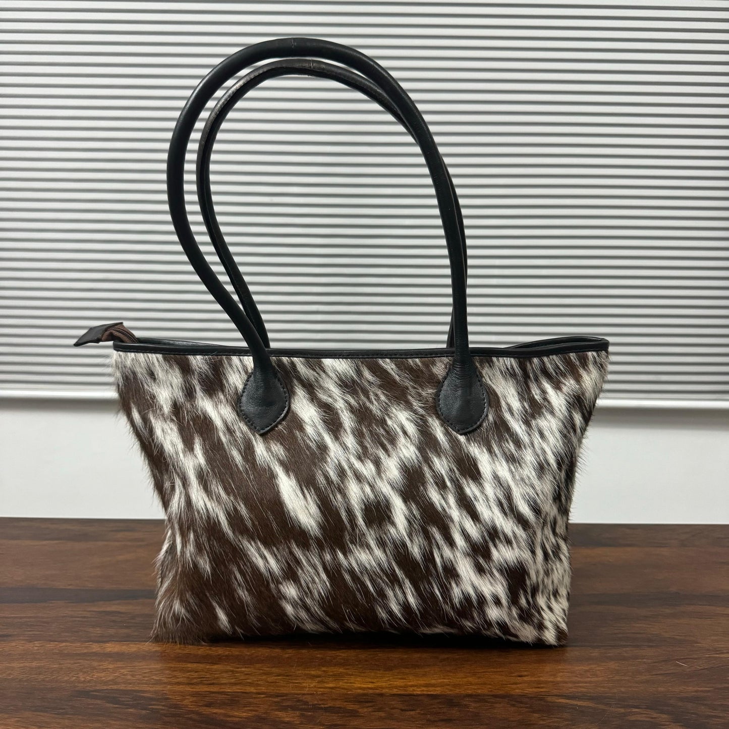 A cowhide handbag with a brown and white mottled pattern and black leather handles, sitting on a wooden table in front of a gray slatted window.