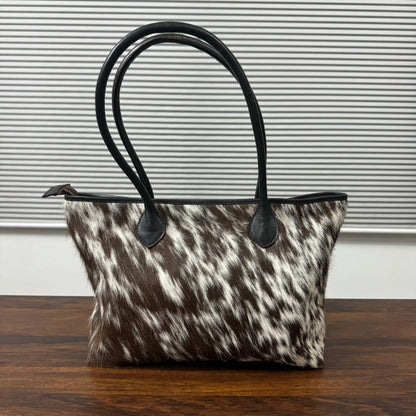 A cowhide handbag with a brown and white mottled pattern and black leather handles, sitting on a wooden table in front of a gray slatted window.