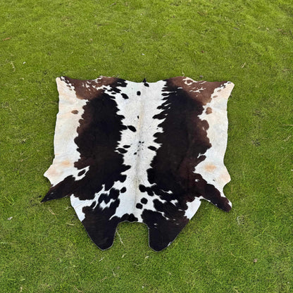 A natural cowhide rug with a black and brown spotted pattern on an off-white base, laid flat on a green grass lawn.