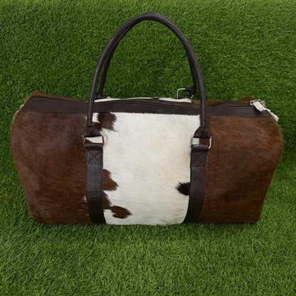 A brown and white cowhide duffel bag with dark brown faux-leather handles and trim, displayed on a green grass background.