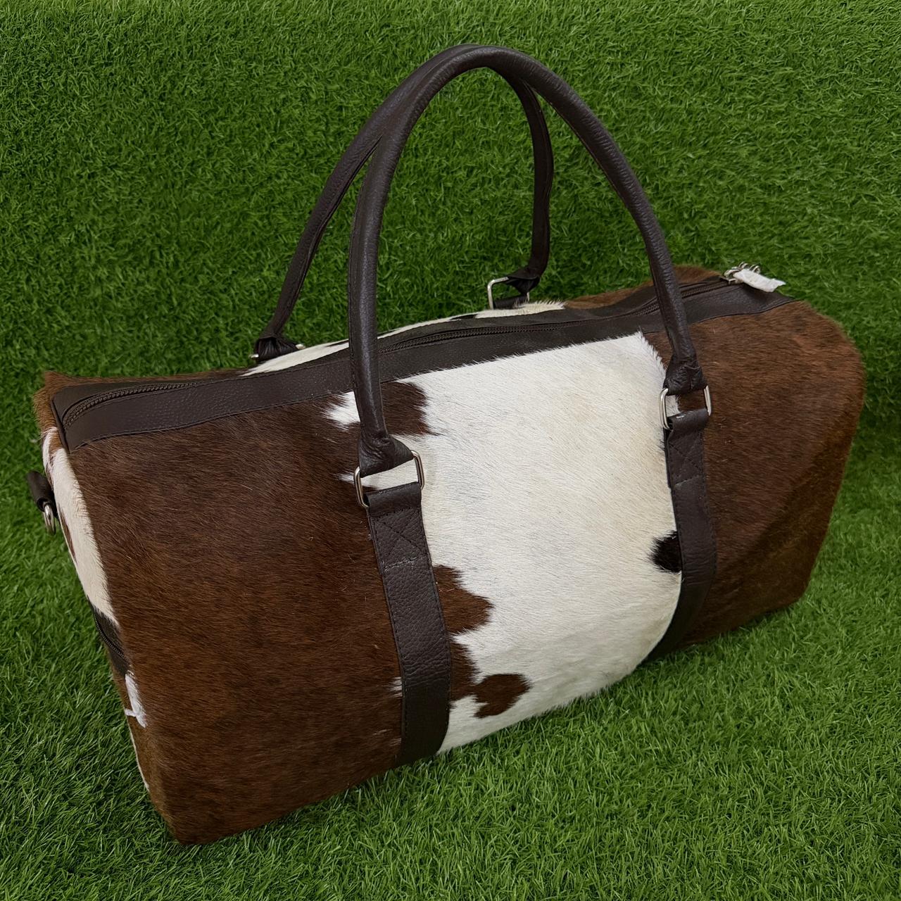 A cowhide duffel bag with brown and white patches, dark brown leather handles and trim, and a matching dark strap, photographed on a green grass-like surface.