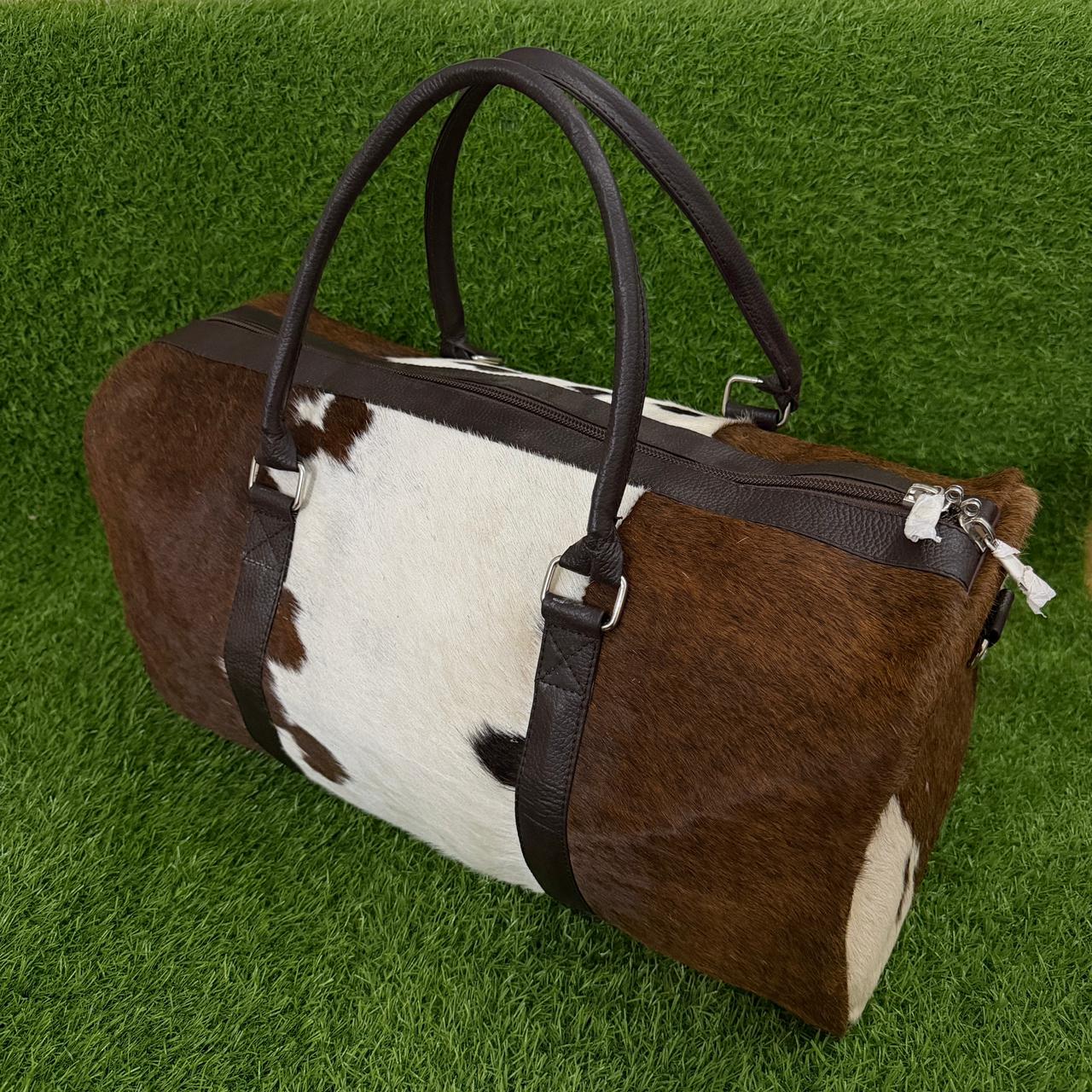 A cowhide duffle bag with brown and white patches and dark brown leather handles and trim, displayed on a green grass background.