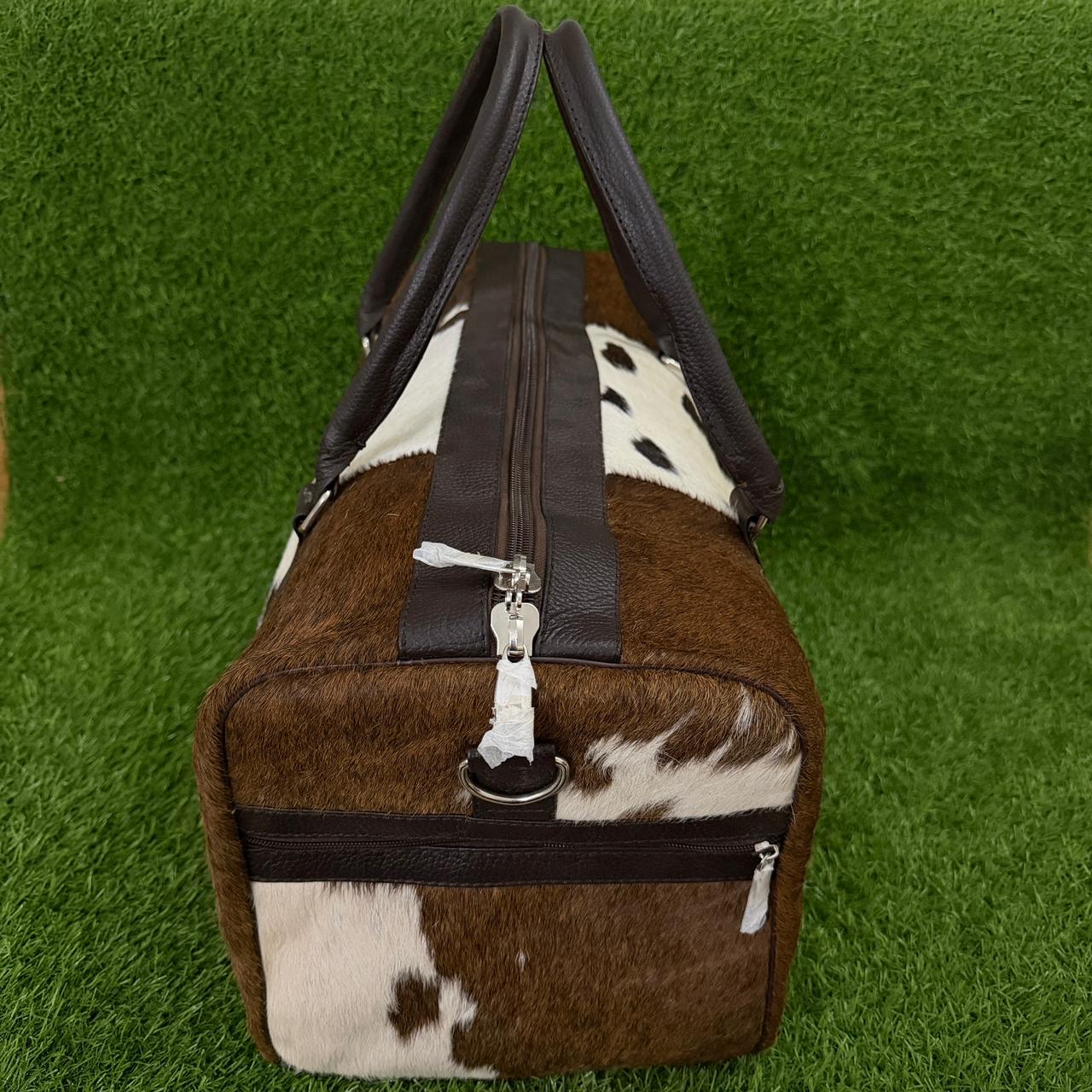 A duffel bag with a brown and white cowhide pattern and dark brown faux-leather trim, shown on a green artificial grass background.