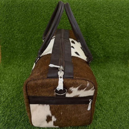 A duffel bag with a brown and white cowhide pattern and dark brown faux-leather trim, shown on a green artificial grass background.
