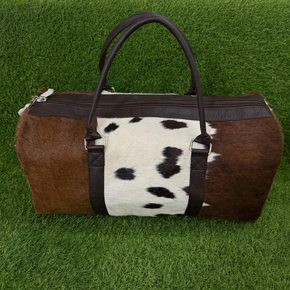 A duffle bag with a brown and white cowhide pattern, dark brown faux-leather handles and trim, and silver zipper hardware, displayed on a green artificial grass background.