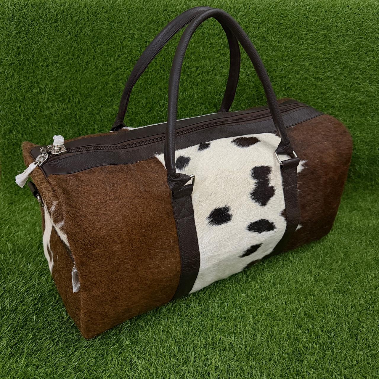 A cowhide duffel bag with brown and black-and-white spotted panels, dark brown handles, and a dark zipper, photographed on a green grass-like surface.