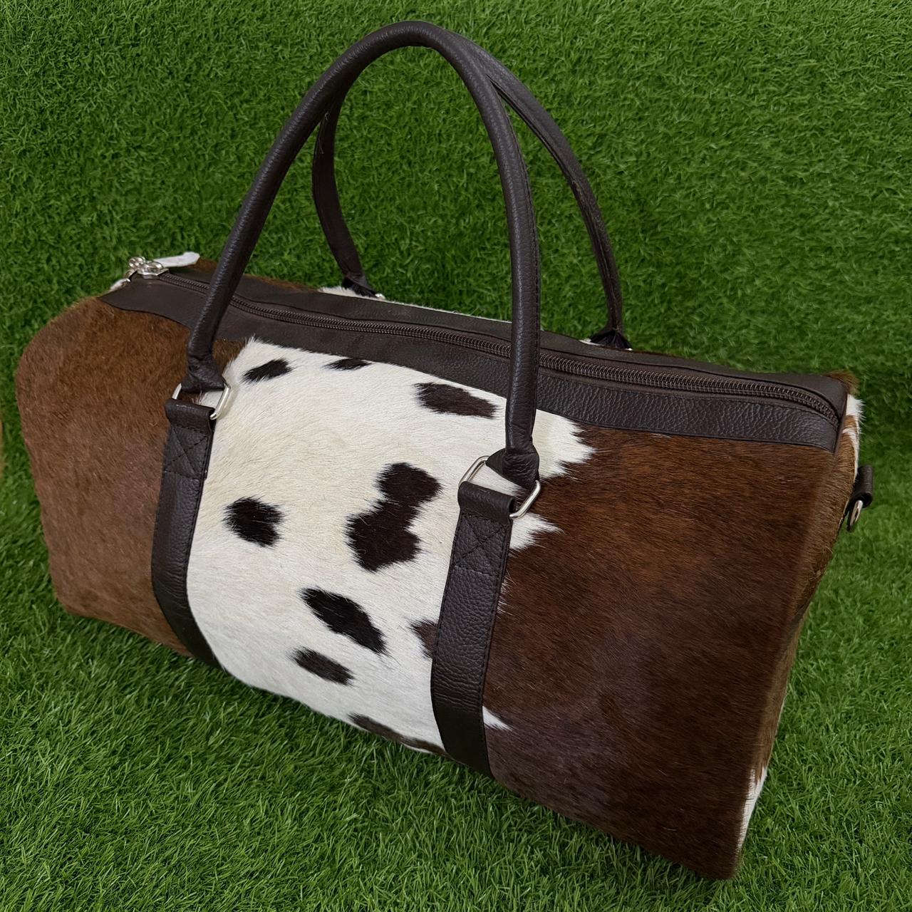 A cylindrical duffle bag with a brown and black cowhide pattern and dark brown faux-leather handles and trim, displayed on a green grass background.