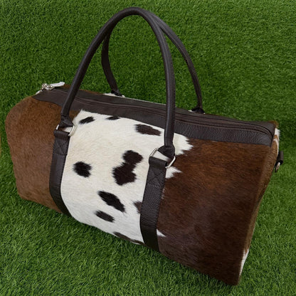A cylindrical duffle bag with a brown and black cowhide pattern and dark brown faux-leather handles and trim, displayed on a green grass background.