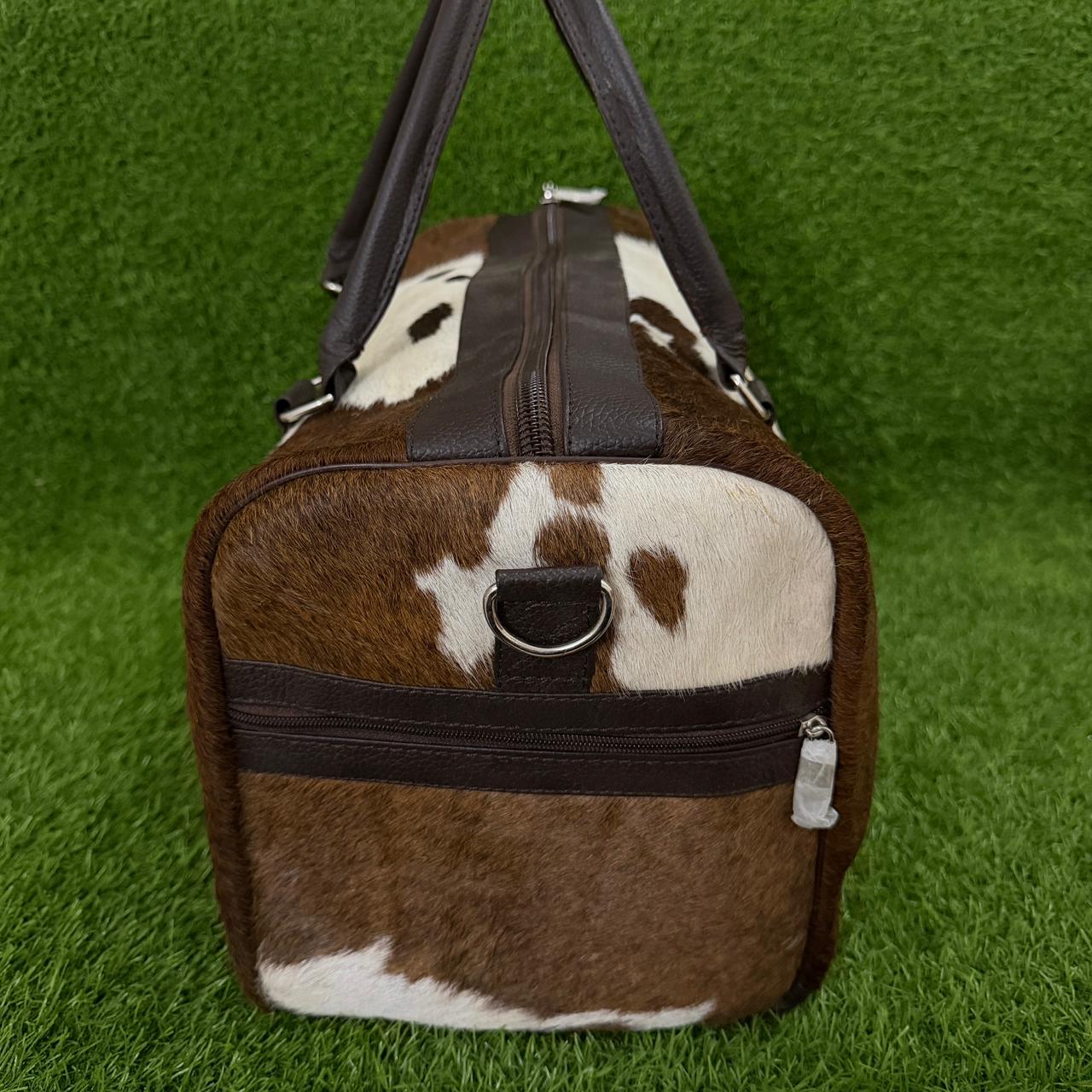A brown and white cowhide duffel bag with dark brown faux-leather trim, shown on a green grass-textured background.
