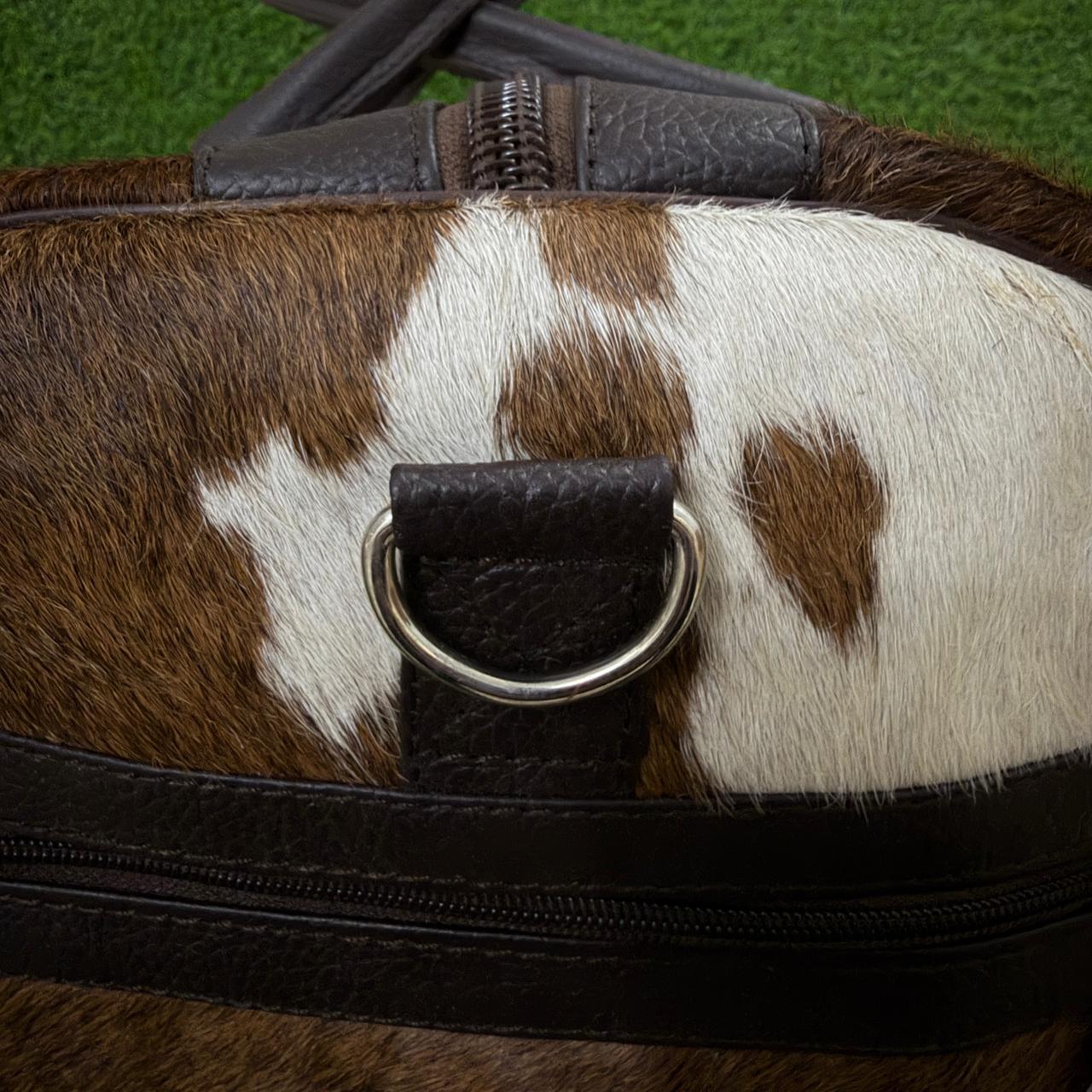 Close-up of a cowhide bag with brown and white spotted pattern, dark brown leather trim, and a silver metal D-ring closure.