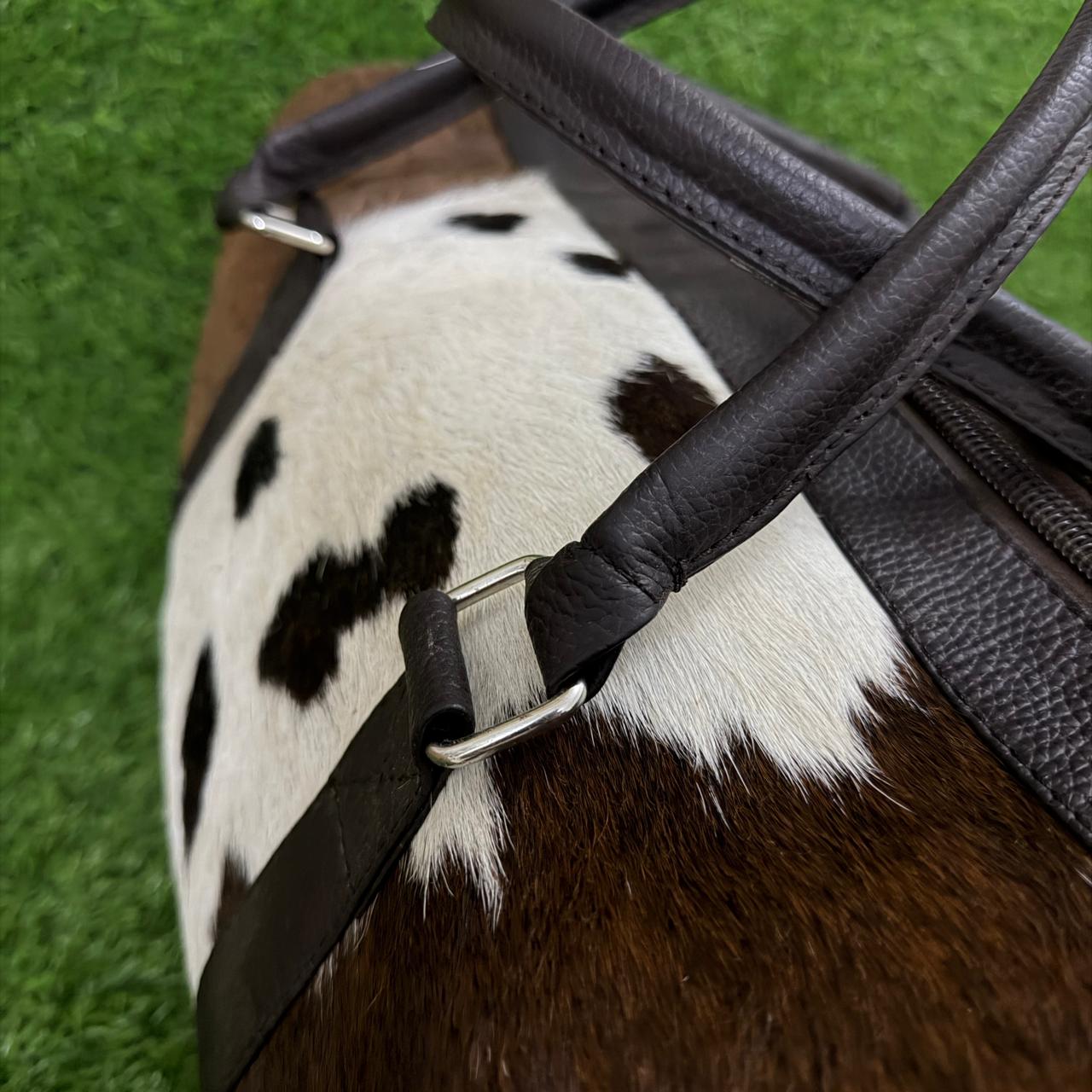 Close-up of a duffel bag with a brown and white cowhide body and dark brown leather handles and trim, shown on a green artificial turf background.