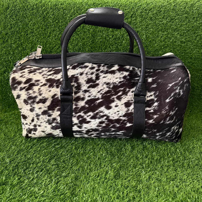 A cowhide-patterned duffel bag with black faux-leather handles and trim, displayed on a green artificial grass background.