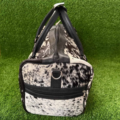 A duffel bag with a black and white cowhide pattern and black faux-leather trim, standing upright on green artificial grass.