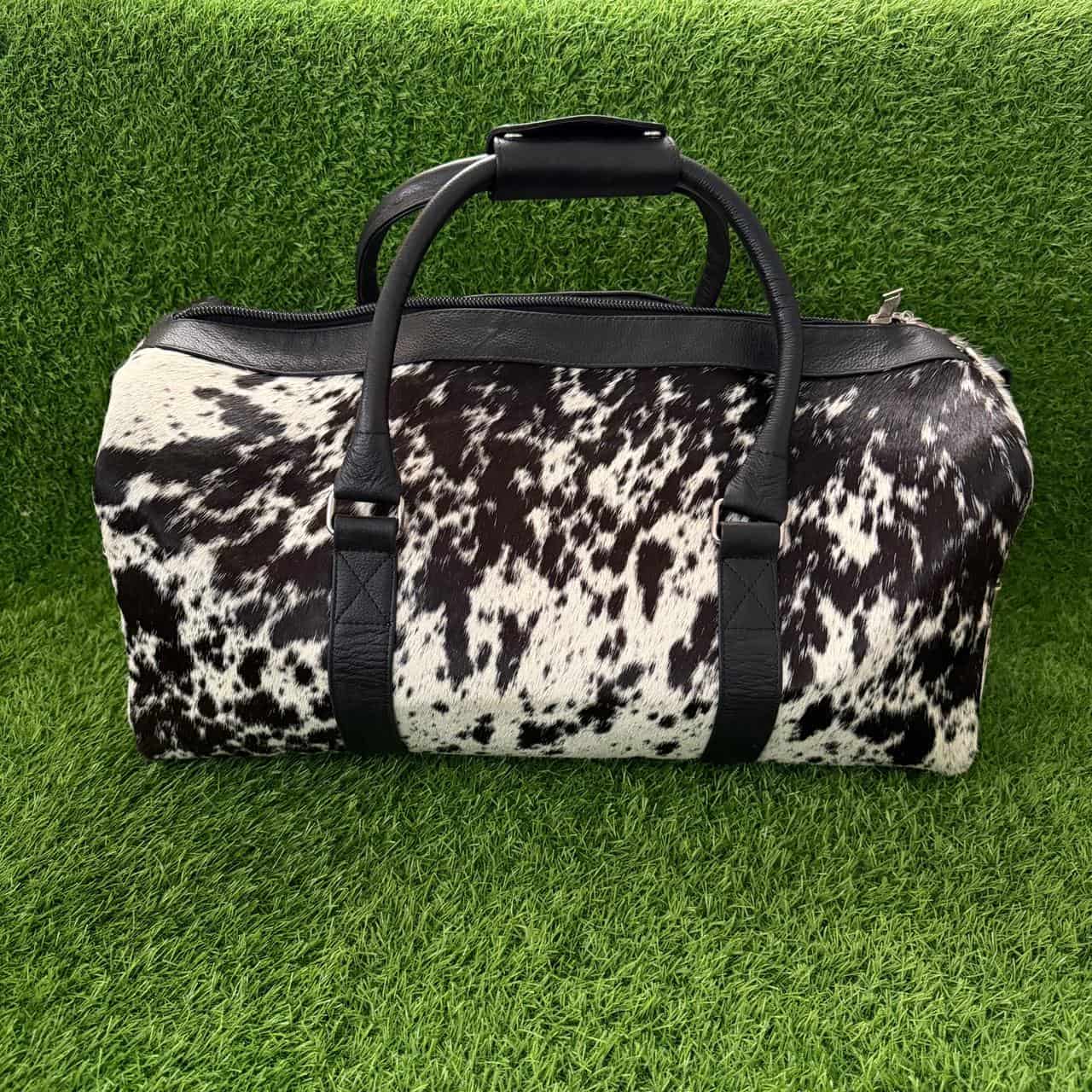 A duffle bag with a black and white cowhide pattern and black faux-leather handles and trim, displayed on a green artificial grass background.