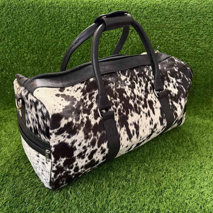 A cowhide-patterned duffel bag with black leather handles and trim, resting on a green grass background.