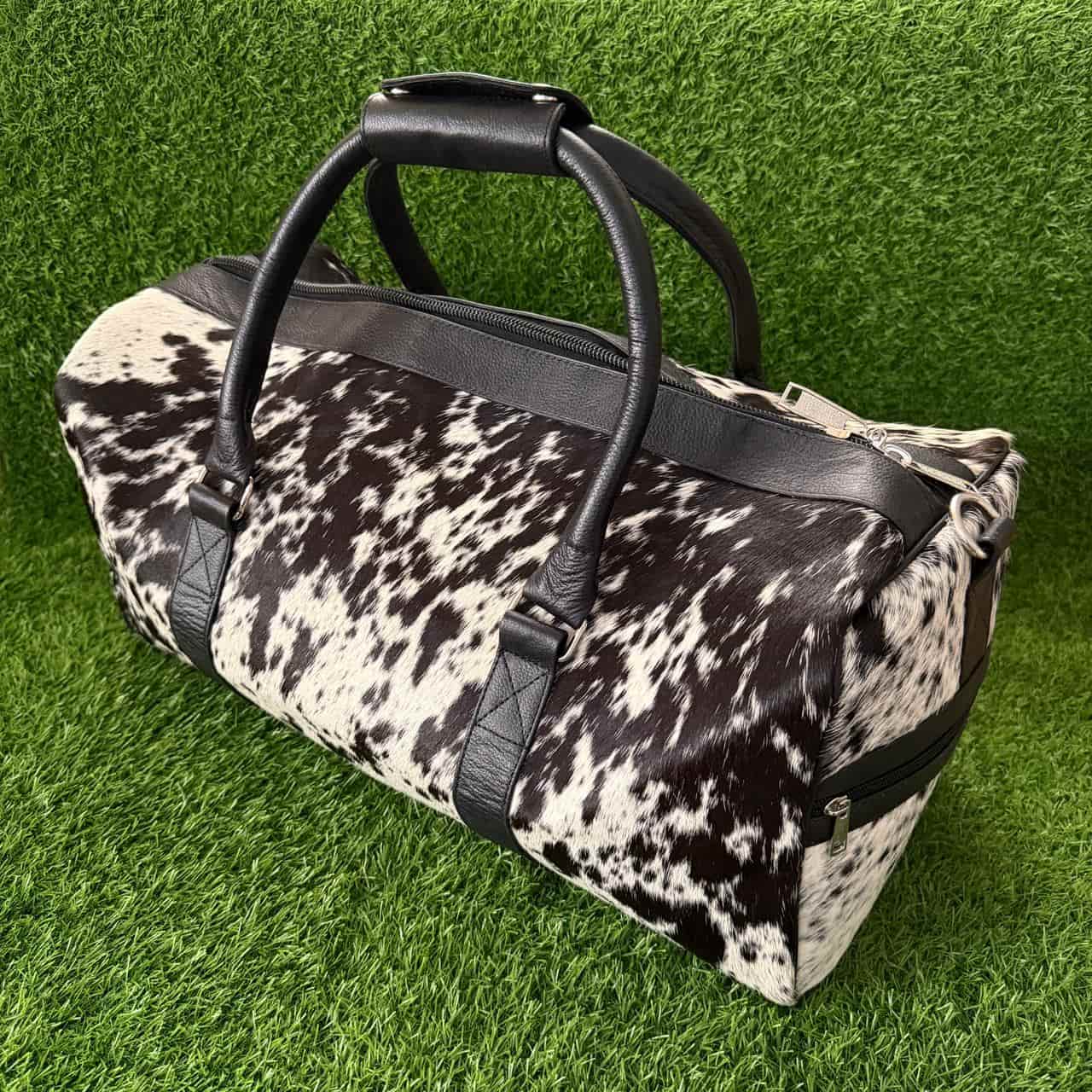A duffle bag with a black and white cow print pattern and black leather handles, displayed on a green artificial grass background.