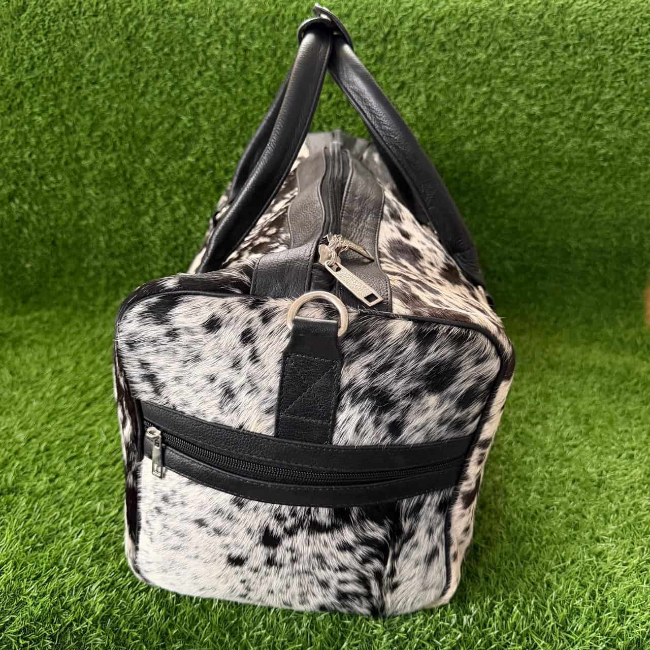 A duffle bag with a black and white cow print pattern and black faux-leather trim, shown on a green artificial grass background.