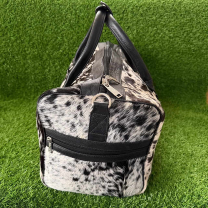 A duffle bag with a black and white cow print pattern and black faux-leather trim, shown on a green artificial grass background.