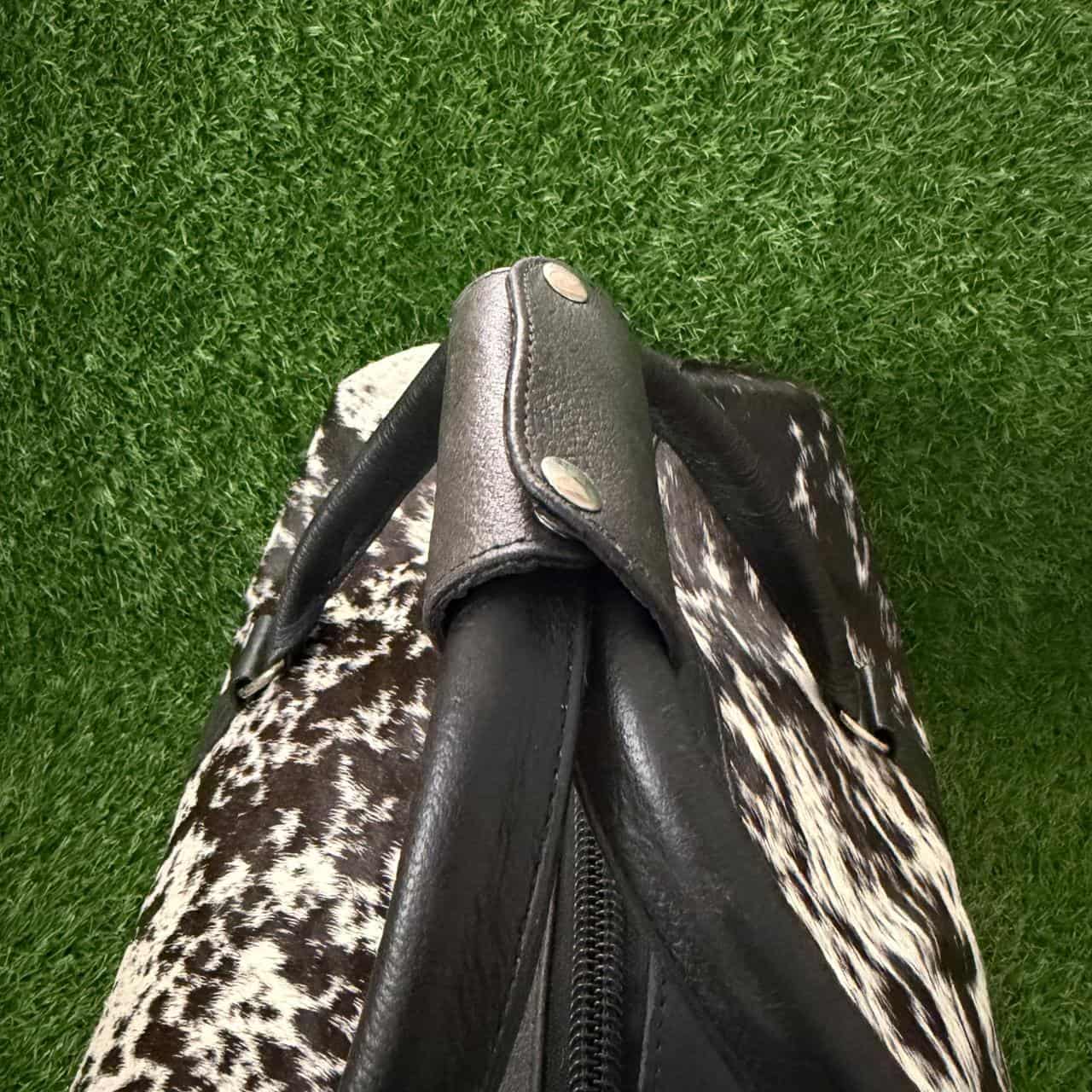 A close-up of a handbag or backpack featuring a black faux-leather strap and zipper trim, attached to a black-and-white cowhide-patterned body, set against a green artificial grass background.