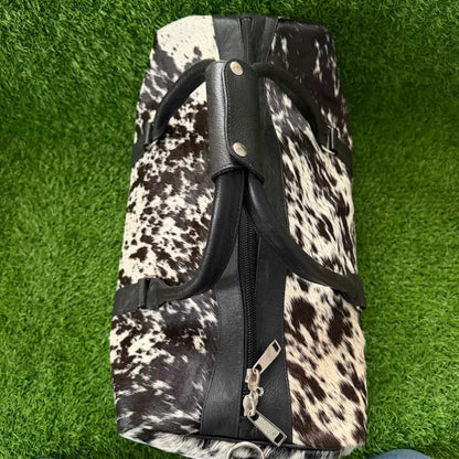 A duffel bag with a black and white cowhide pattern and black faux-leather trim, shown from a top-down view on green artificial grass.