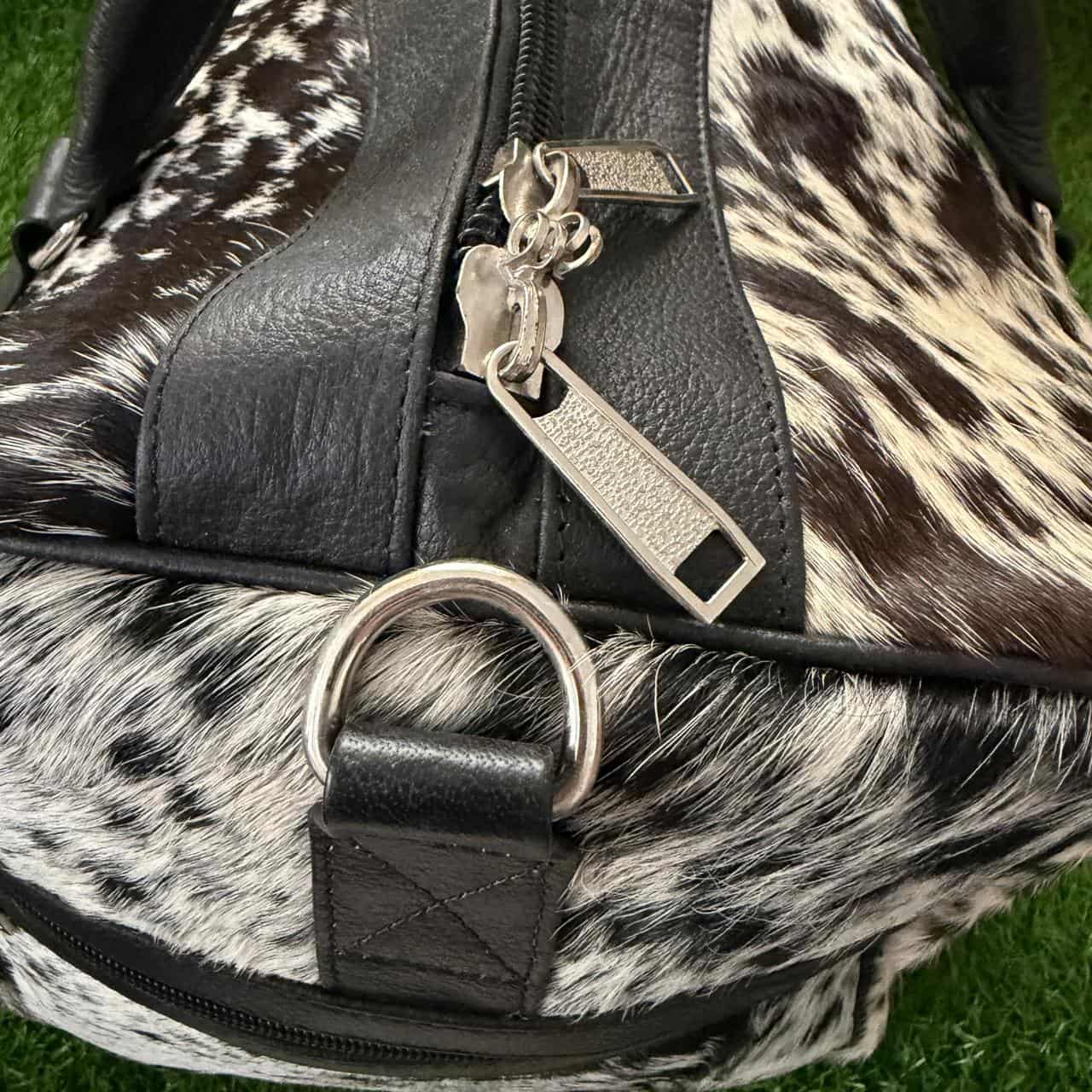 Close-up of a cowhide and black faux-leather bag with a silver zipper pull and metal D-ring, resting on green grass.