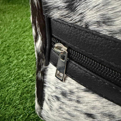 Close-up of a black faux-leather zipper trim with a silver metal pull on a cowhide bag, set against green artificial grass.