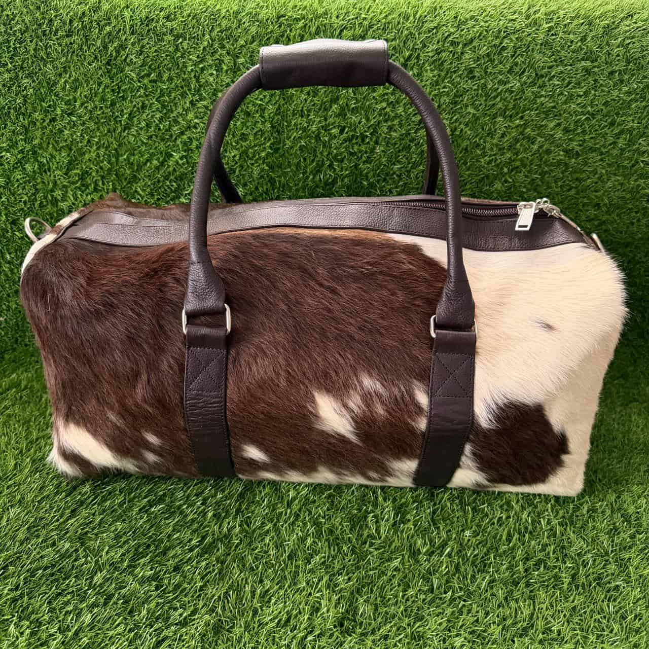 A duffel bag made from brown and white cowhide with dark brown leather handles and trim, photographed on a green grass-like surface.
