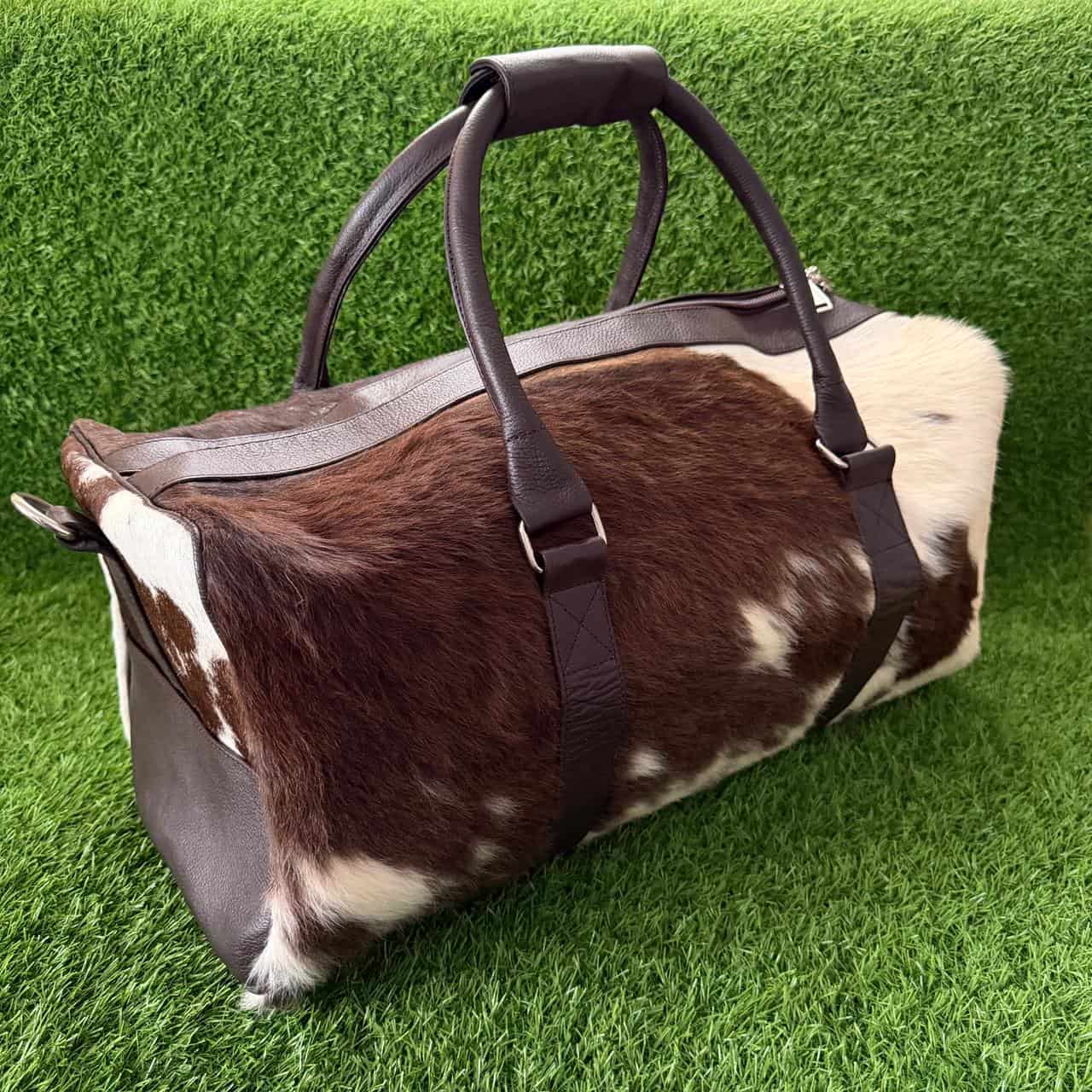 A duffel bag made from brown and white cowhide with dark brown leather handles and trim, photographed on a green grass-like surface.
