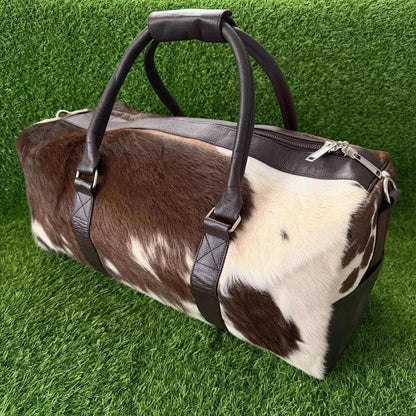 A duffel bag made from brown and white cowhide with dark brown leather handles and trim, photographed on a green grass-like surface.