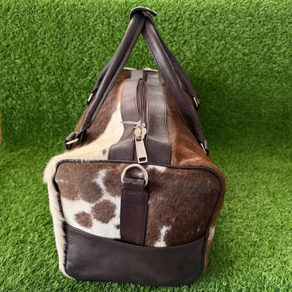 A duffel bag with a brown and white cowhide pattern and dark brown leather trim, shown on a green artificial grass background.