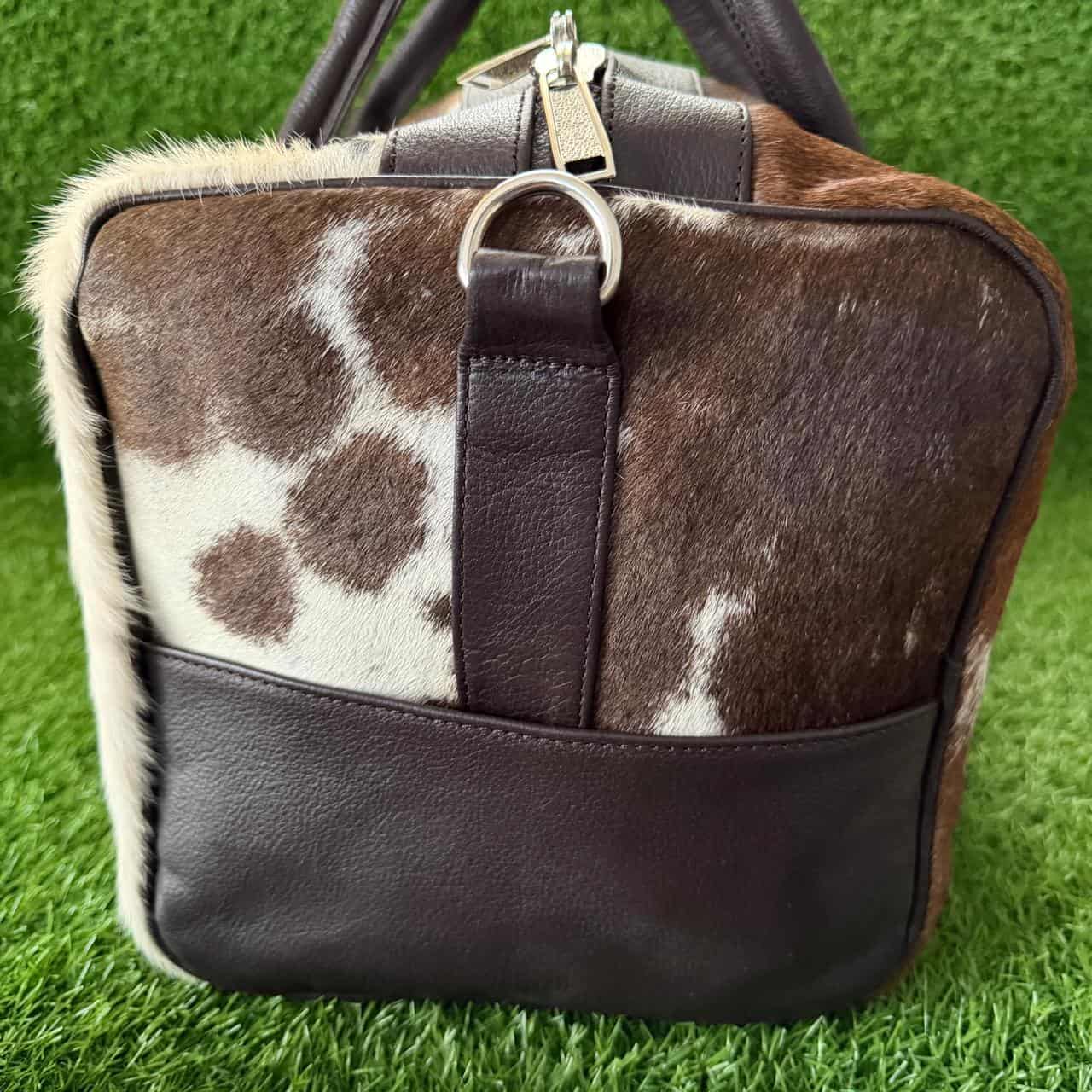 A structured handbag combining cowhide and dark brown leather, featuring a brown and white spotted cowhide body, dark brown leather trim, handles, and a bottom panel, set against a green grass background.