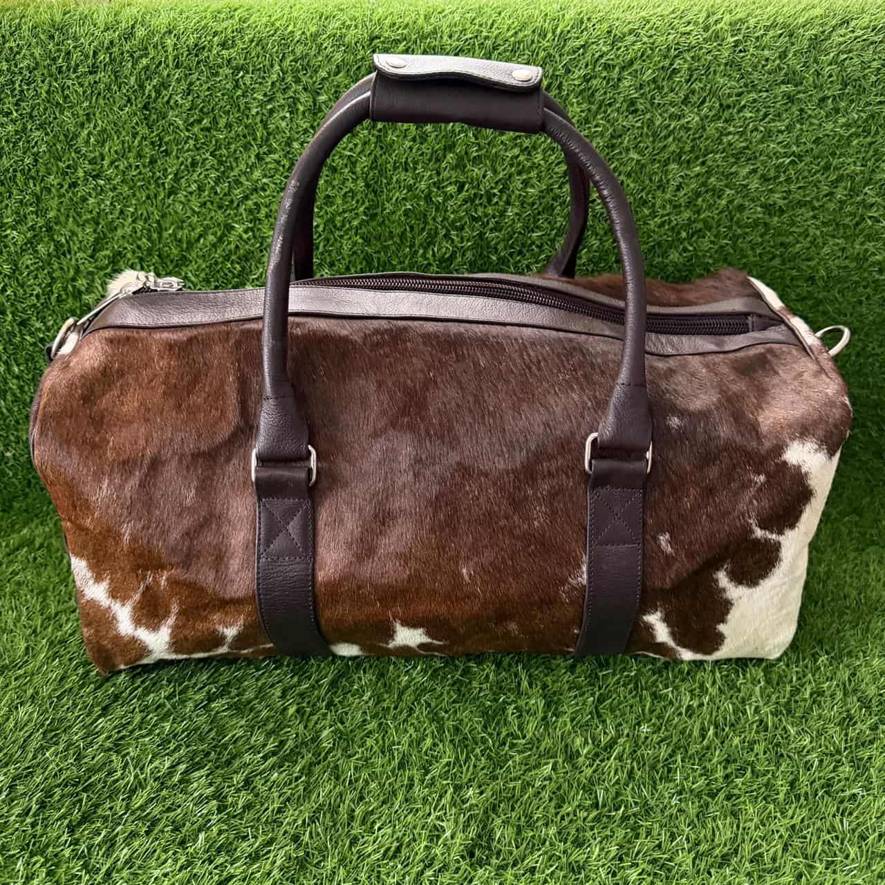 A brown and white cowhide duffel bag with dark brown leather handles and straps, displayed on a green artificial grass background.
