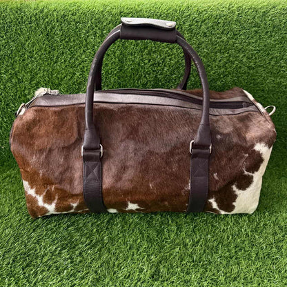 A brown and white cowhide duffel bag with dark brown leather handles and straps, displayed on a green artificial grass background.