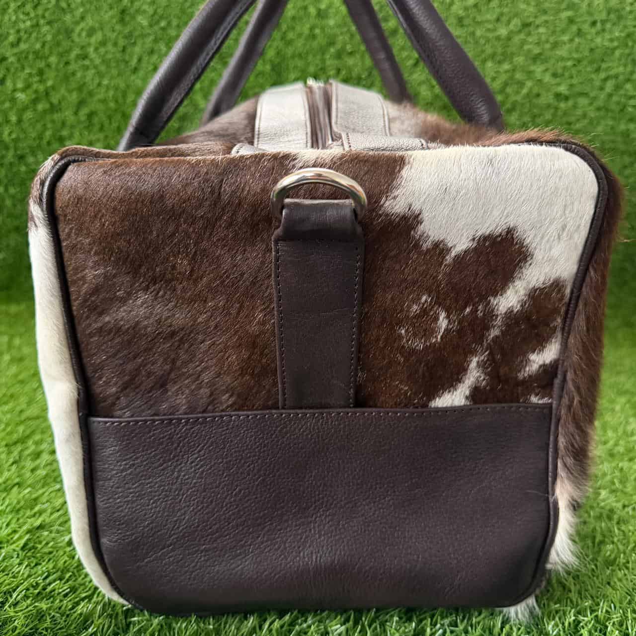 A cowhide and dark brown leather duffel bag with dark brown handles, shown on a green grass-like background.