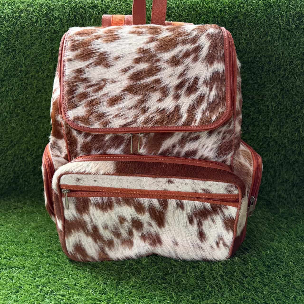 A backpack with a brown and white cow print pattern and reddish-brown trim, shown against a green grass-like background.