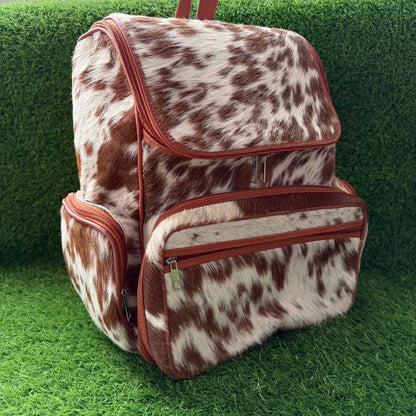 A cowhide-patterned backpack with reddish-brown trim and zipper details, shown on a green grass-like surface.
