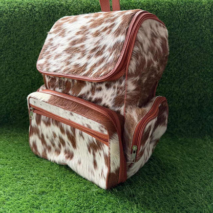 A backpack and matching zippered pouch set in brown and white cowhide with tan leather trim, displayed on artificial green grass.