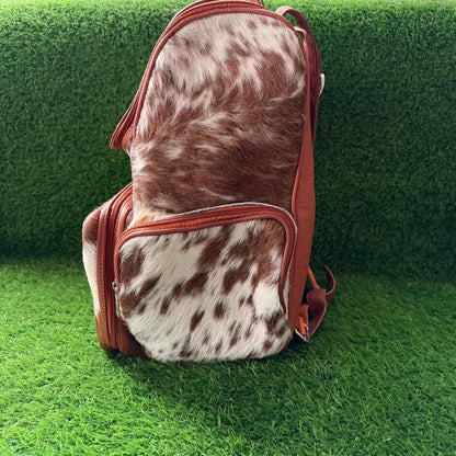 A small backpack with a brown and white cowhide pattern and reddish-brown leather trim, standing on green artificial grass against a matching green background.