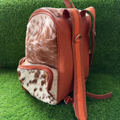 A brown leather and cowhide backpack with a matching brown shoulder strap, shown on a green artificial grass background.