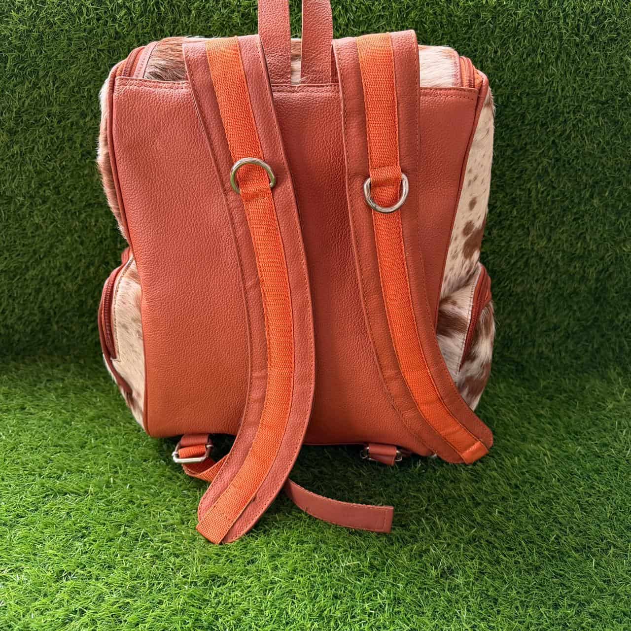 A backpack with a textured terracotta orange leather front and back, accented with brown and white cowhide side panels, photographed on a green artificial grass background.