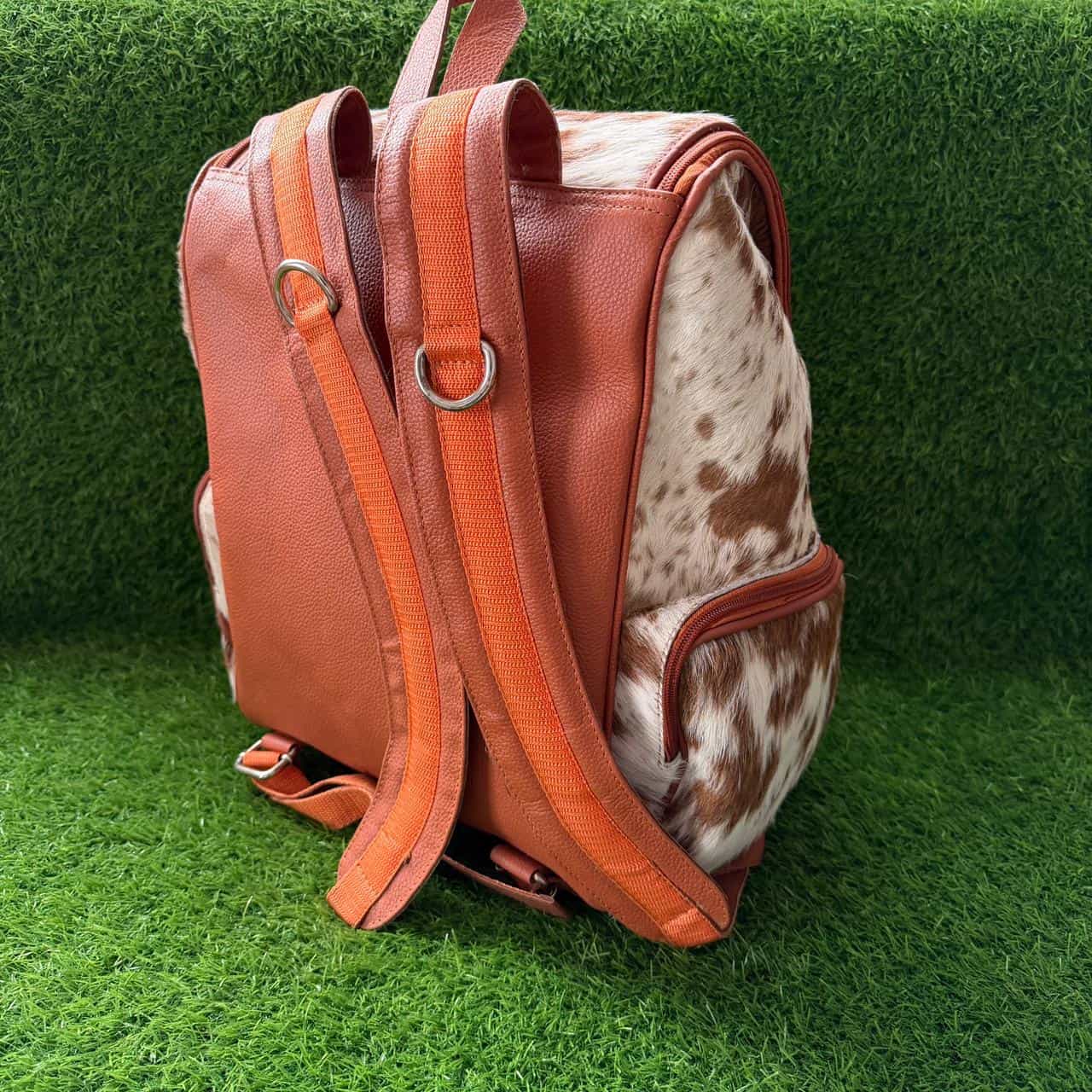 A brown leather and cowhide backpack standing upright on artificial green grass.