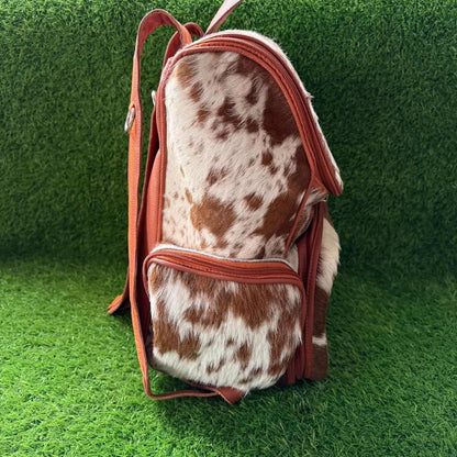 A small backpack made from cowhide with a brown and white spotted pattern, featuring rust-brown leather trim and straps, set against a green artificial grass background.