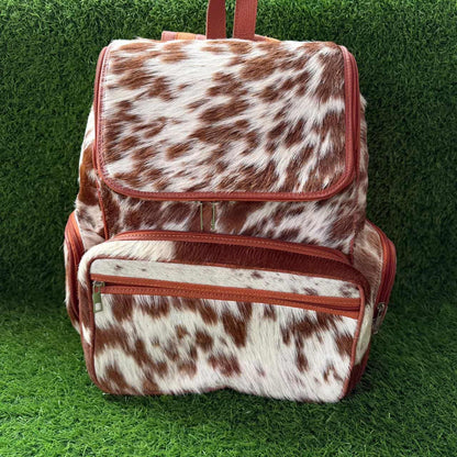A backpack with a brown and white cowhide pattern and reddish-brown trim, standing on a green grass-like surface against a matching green background.