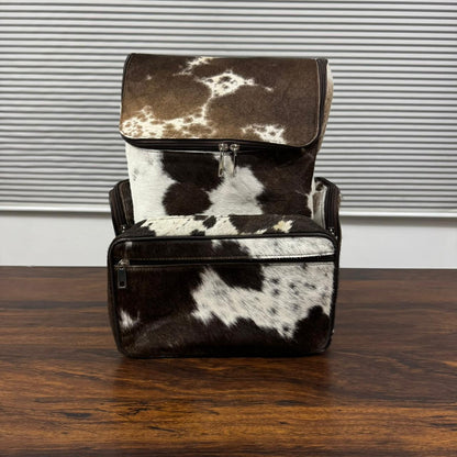 A cowhide backpack with a brown and white spotted pattern, standing on a dark wooden table in front of a gray slatted wall.