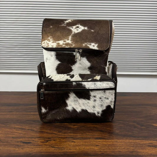 A cowhide backpack with a brown and white spotted pattern, standing on a dark wooden table in front of a gray slatted wall.
