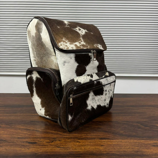 A cowhide backpack and matching pouch set with a brown and white spotted pattern, displayed on a dark wooden table against a gray slatted background.