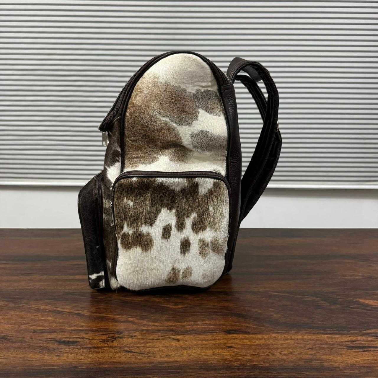 A small shoulder bag with a brown and white cowhide pattern and black trim, standing on a dark wooden table in front of a window with horizontal blinds.