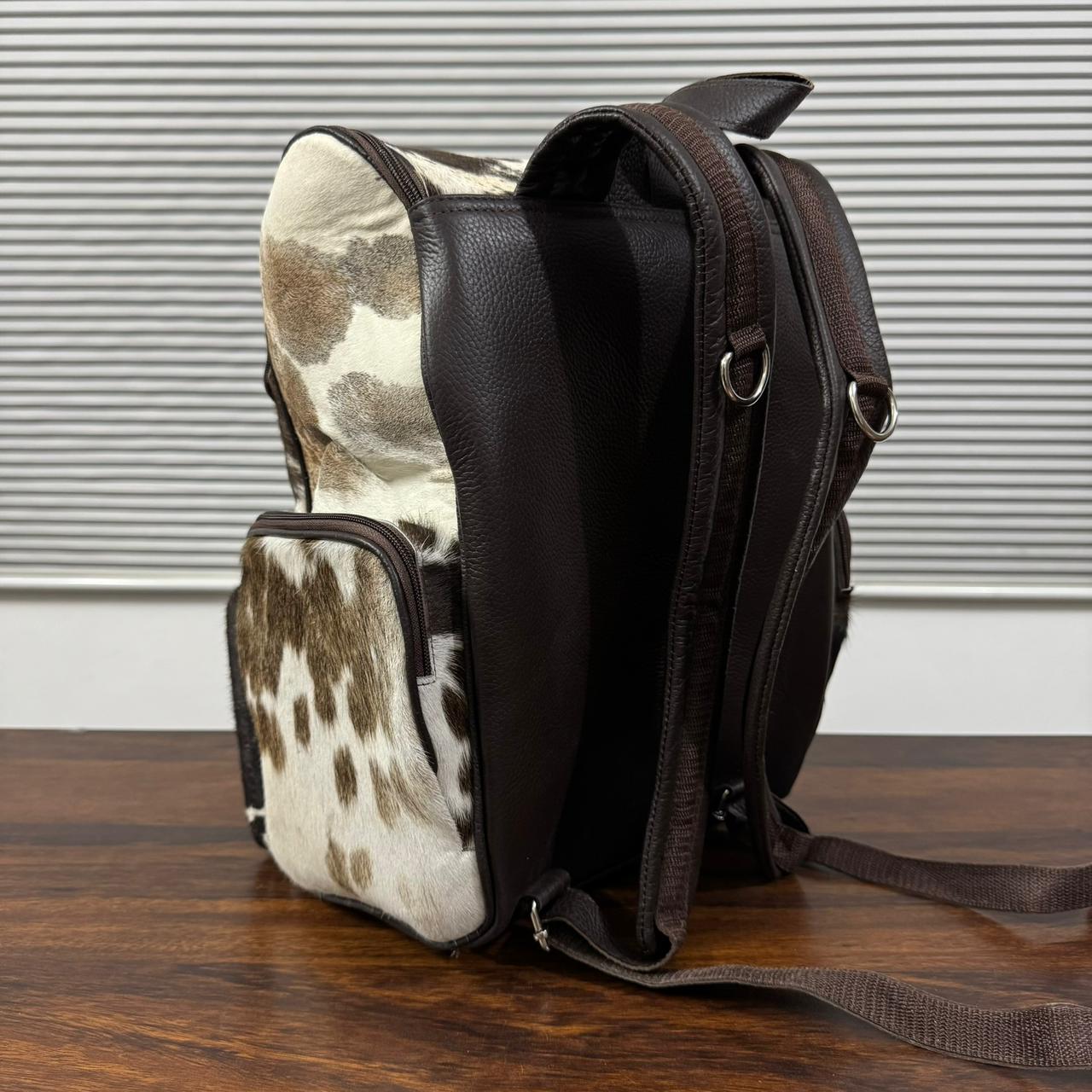 A brown and cowhide-patterned shoulder bag standing on a wooden tabletop in front of a gray slatted window.